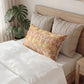 A decorative lumbar pillow with an orange, yellow, and white floral pattern rests on a bed with white bedding and beige pillows, next to a potted monstera plant.