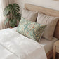 A styled bedroom scene showing a bed with white bedding, two beige quilted pillows, and a decorative rectangular lumbar pillow with a floral and scroll pattern in sage green, beige, and muted orange. A potted monstera plant sits beside the bed.