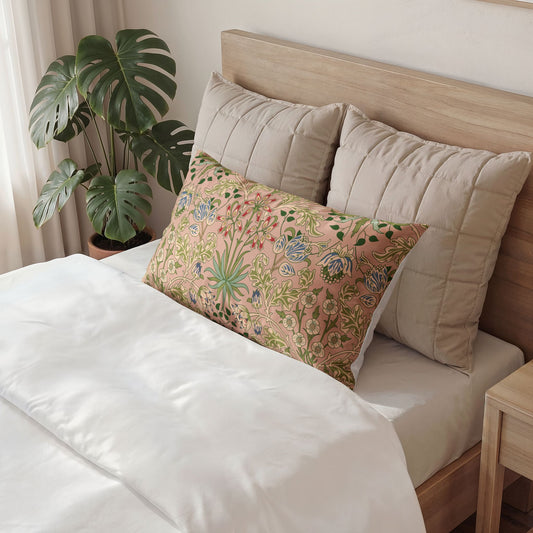 A decorative rectangular throw pillow with a colorful botanical pattern in pink, green, and blue, styled on a bed with white bedding and beige pillows next to a potted monstera plant.