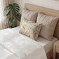 A styled bed with white bedding, two large beige quilted pillows, and a smaller decorative throw pillow featuring a vintage-style bird and floral pattern. A wooden headboard and a potted monstera plant are visible in a bright bedroom setting.