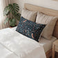 A dark navy decorative pillow with a detailed botanical and bird pattern, styled on a bed with beige pillows and white bedding next to a potted monstera plant.
