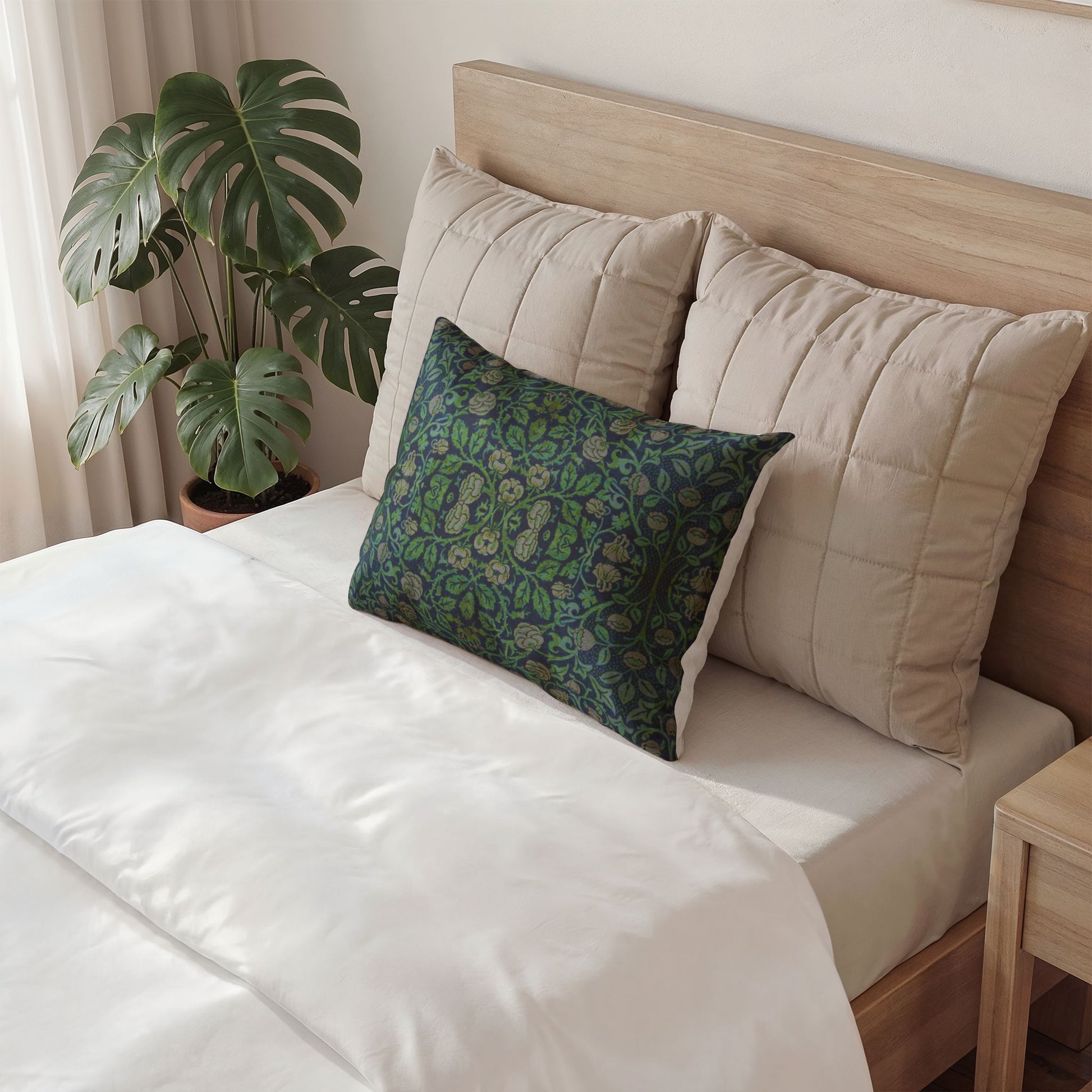 A styled bed with white bedding, two large beige quilted pillows, and a decorative square throw pillow featuring a green and blue paisley pattern. A wooden headboard and a potted monstera plant are visible in a bright bedroom setting.