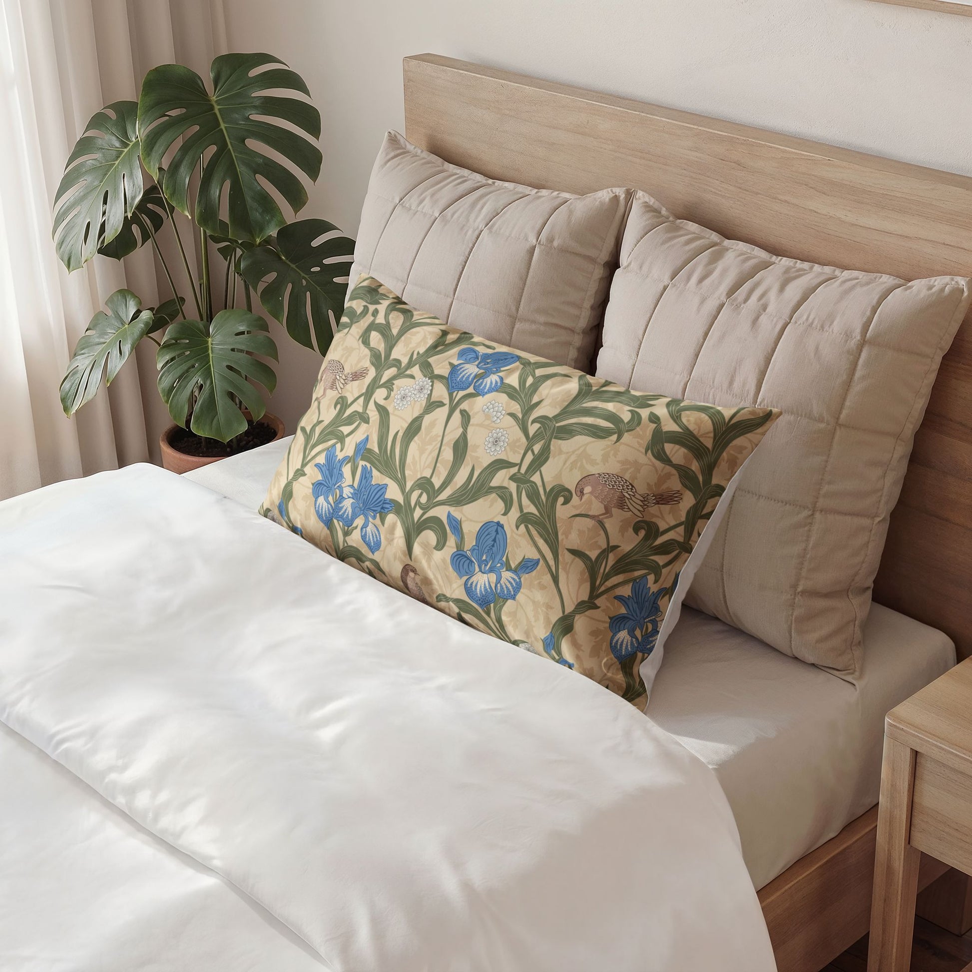 A decorative rectangular throw pillow with a blue and green floral pattern rests on a bed with white bedding and beige pillows, next to a potted monstera plant.