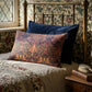 A decorative rectangular lumbar pillow with an intricate botanical and floral pattern in orange, green, and purple, styled on a bed with a dark navy blue pillow and a brass headboard in a vintage bedroom setting.
