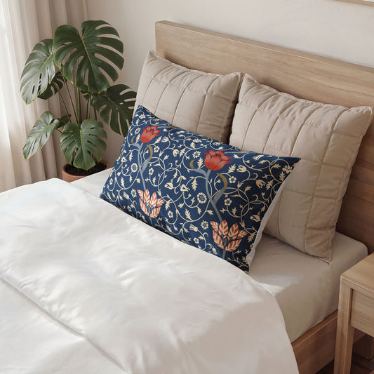 A navy blue decorative pillow with a floral and vine pattern in orange, beige, and green, styled on a bed with white bedding and two beige pillows next to a wooden headboard and a potted monstera plant.