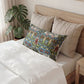 A decorative rectangular lumbar pillow with a colorful floral pattern in blue, green, and red, styled on a bed with beige pillows and white bedding in a bedroom setting.