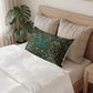 A decorative rectangular lumbar pillow with a detailed botanical and bird pattern in dark green, blue, and orange, styled on a bed with white bedding and beige pillows in a bedroom setting.