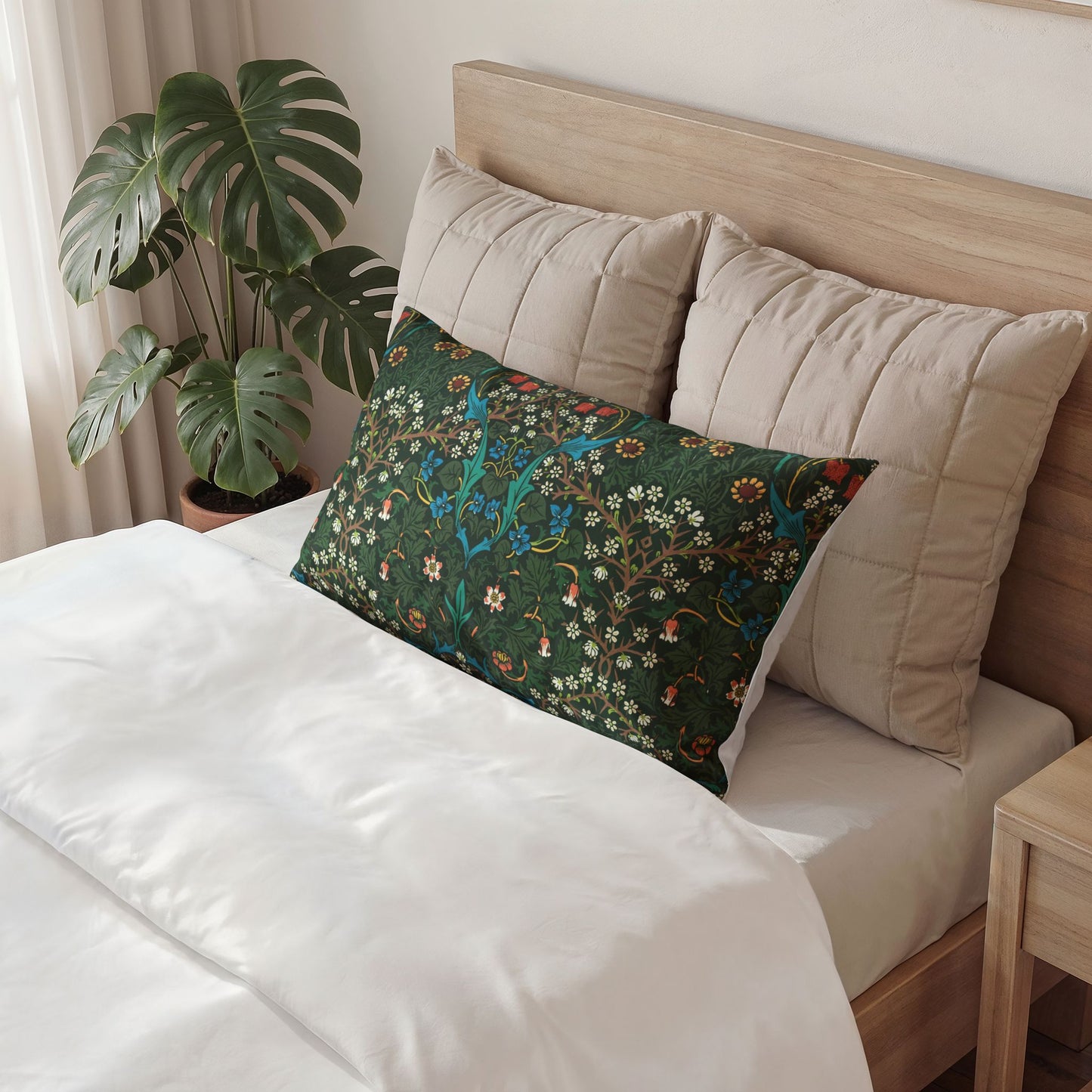 A decorative rectangular lumbar pillow with a detailed botanical and bird pattern in dark green, blue, and orange, styled on a bed with white bedding and beige pillows in a bedroom setting.