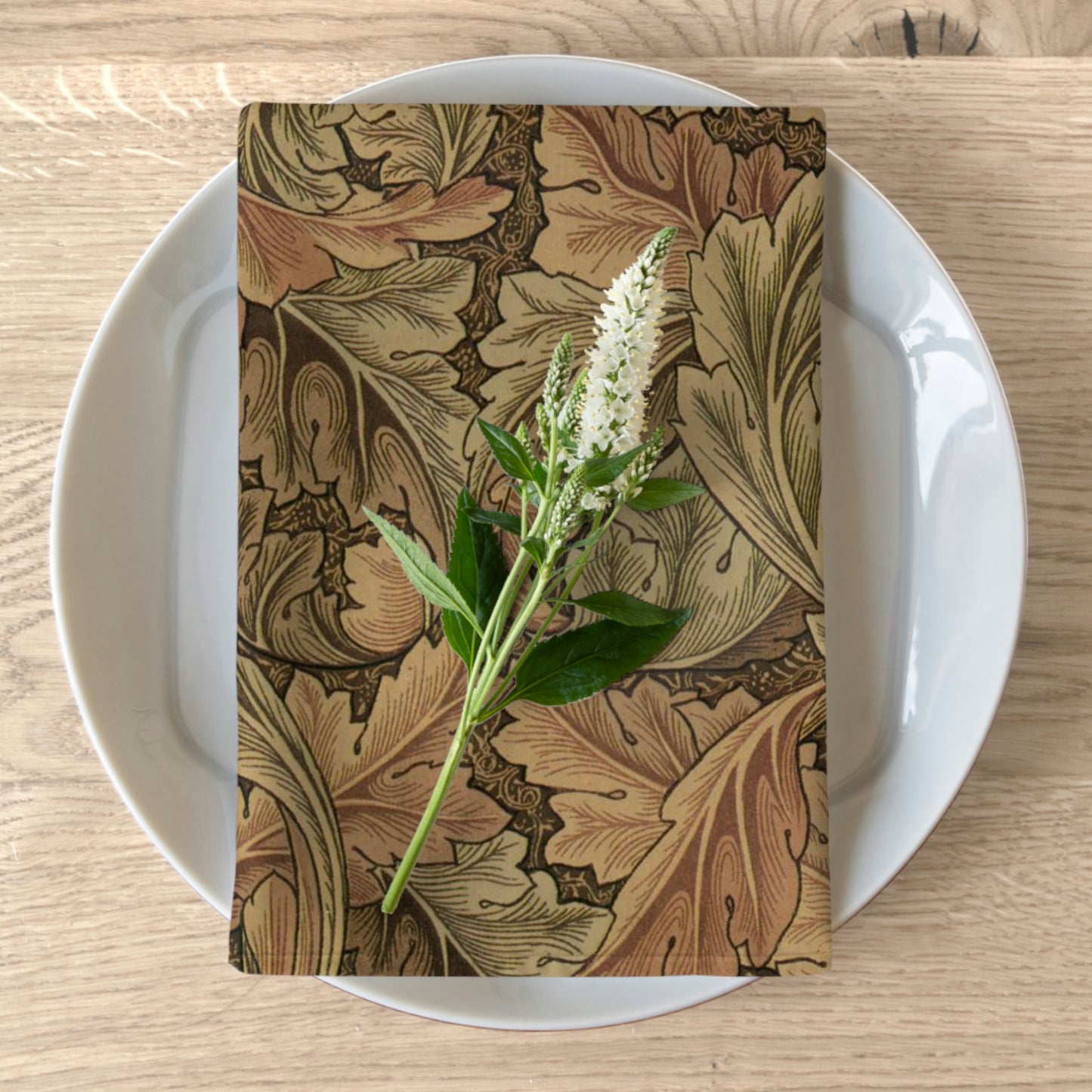 A brown cotton twill napkin with a floral design, inspired by William Morris, placed on a plate on a wooden table.