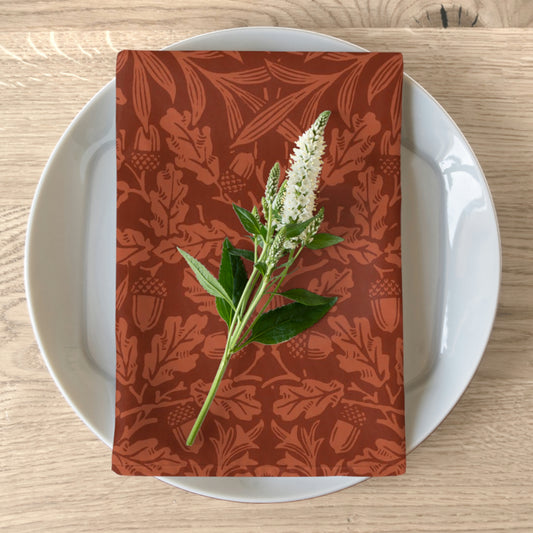 A rust-coloured cotton twill napkin with a floral and acorn pattern, displayed on a plate with a floral arrangement on top.