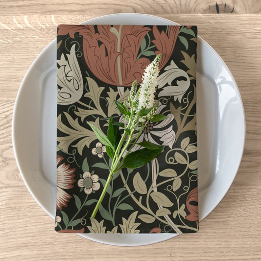 A floral patterned cotton twill napkin inspired by William Morris, displayed on a white plate with a piece of floral decor to the napkin's left.
