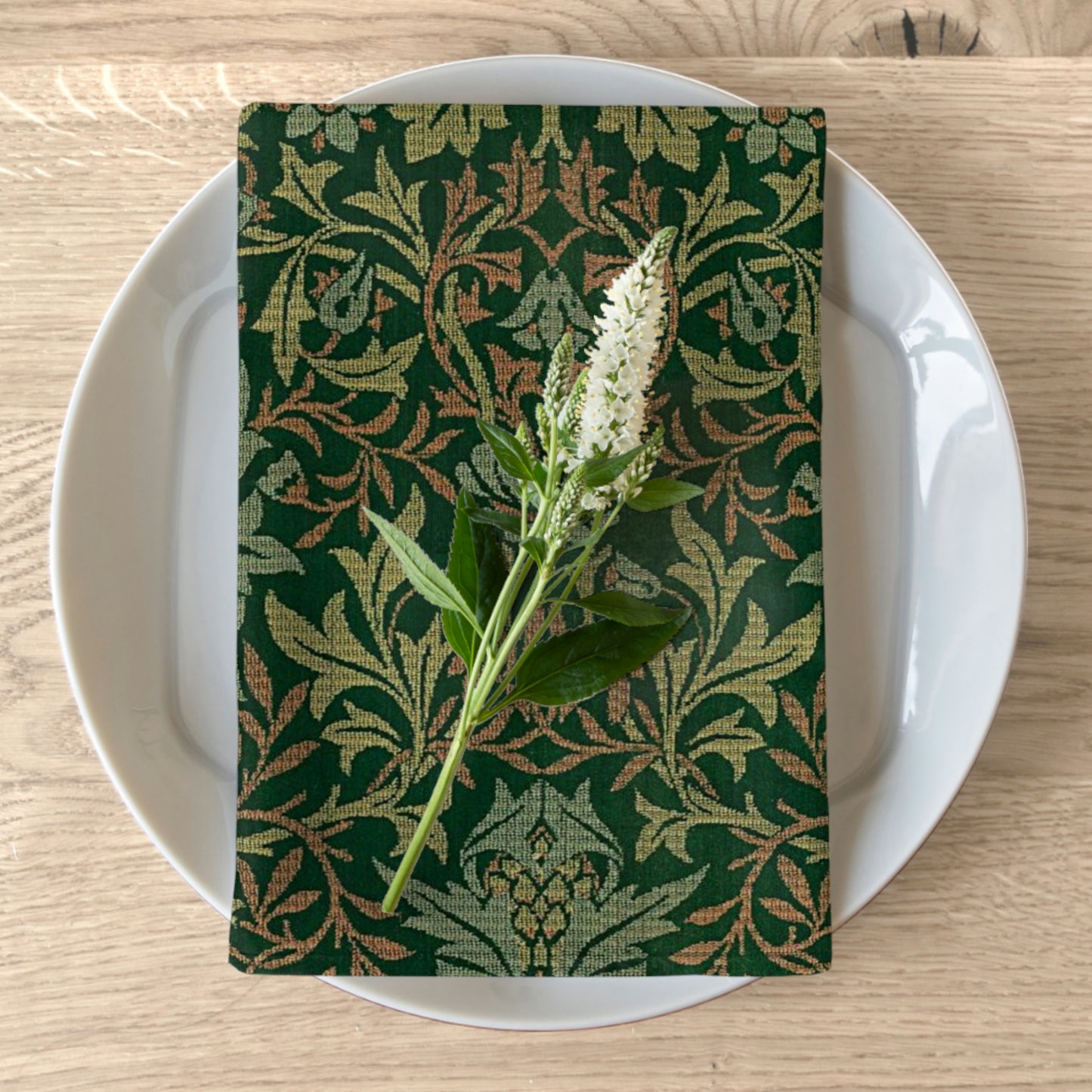 A green floral patterned cotton twill napkin is displayed on a white plate with a piece of greenery on top, set against a wooden table background.