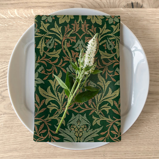 A green floral patterned cotton twill napkin is displayed on a white plate with a piece of greenery on top, set against a wooden table background.