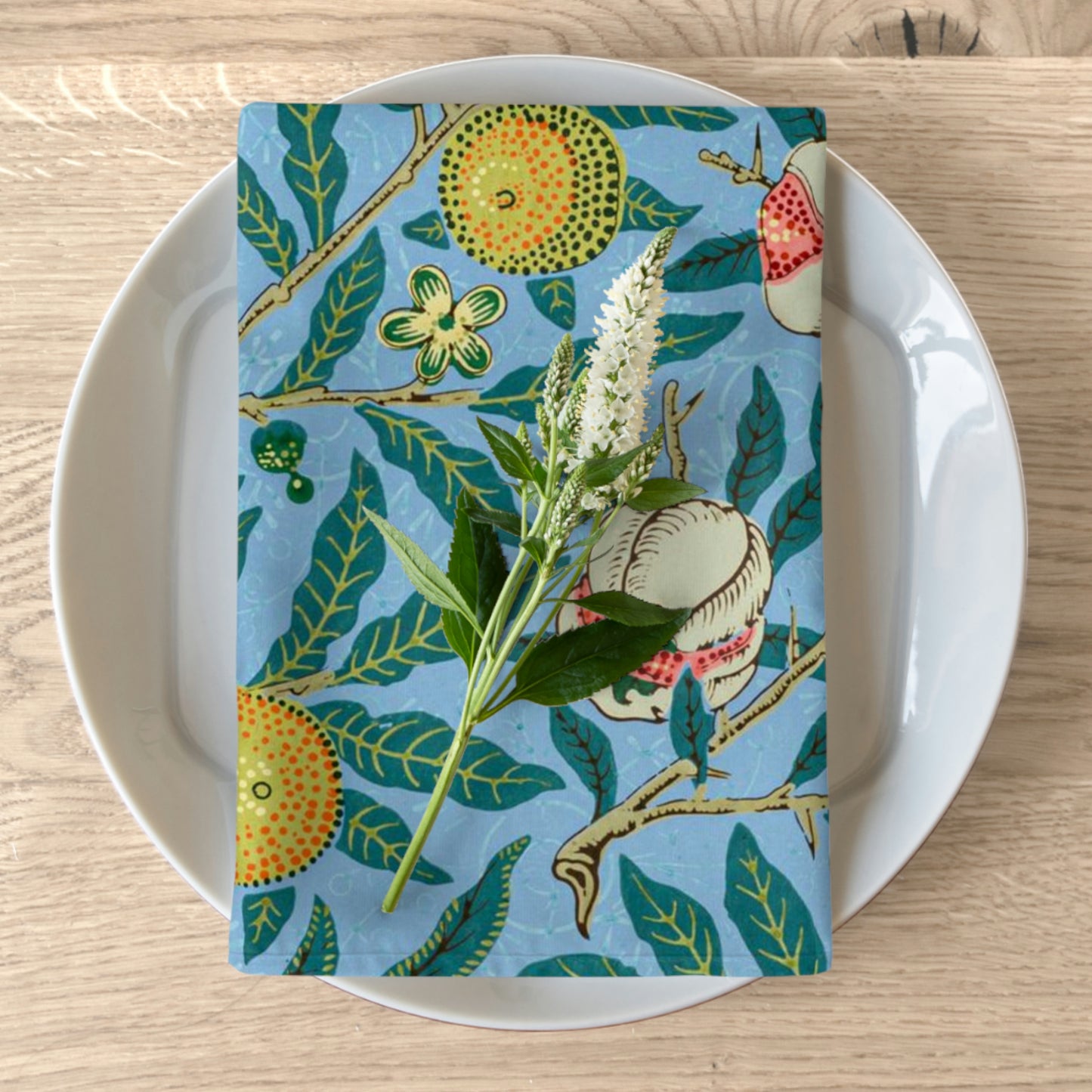 A blue cotton twill napkin with a floral design featuring fruits and leaves, placed on a plate on a wooden table.