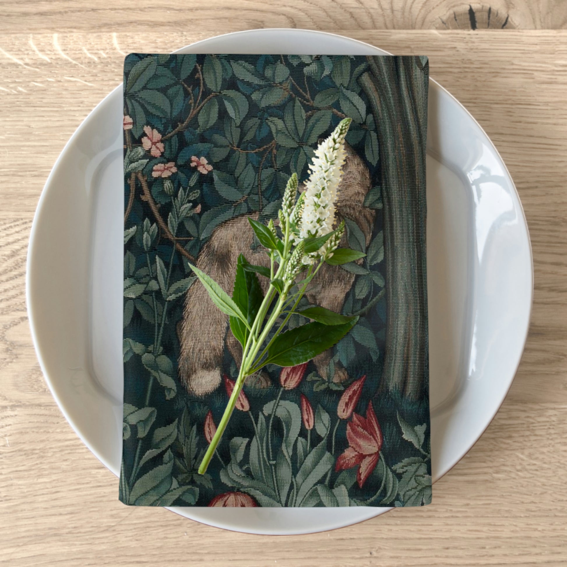 A cotton twill napkin with a floral and animal print, likely inspired by William Morris, displayed on a plate with a piece of greenery placed on top.