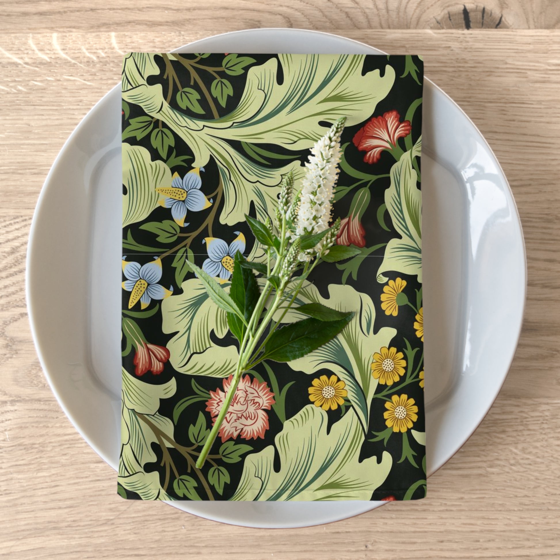 A green cotton twill napkin with a floral design by William Morris, placed on a plate on a wooden table.
