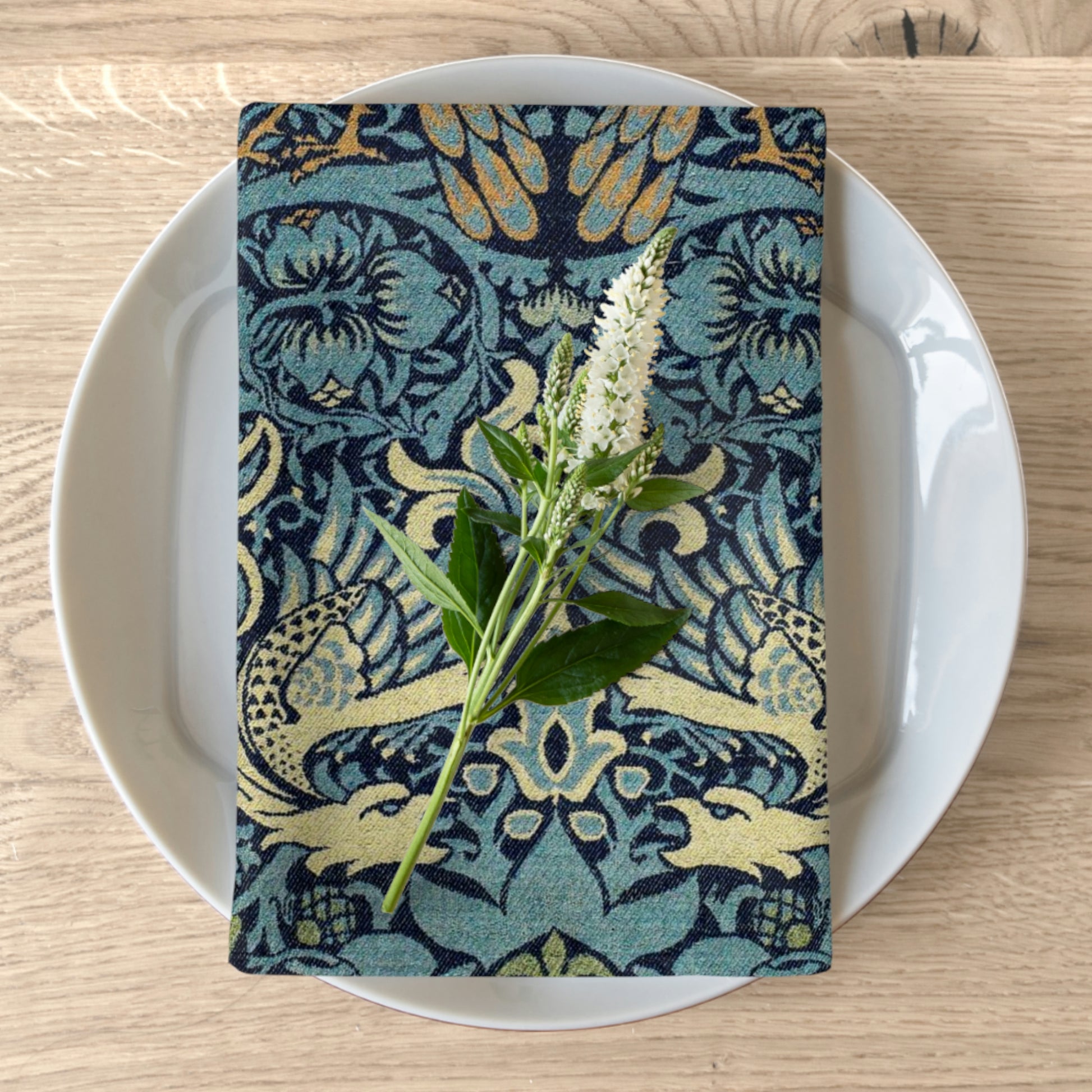A floral printed cotton twill napkin, in shades of blue and green, is placed on a white plate with a piece of greenery on top, on a wooden table.