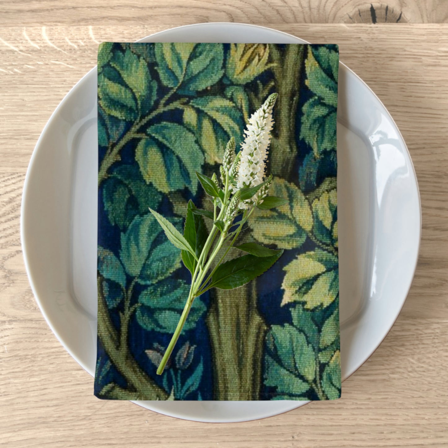 A green cotton twill napkin with a floral design, placed on a plate on a wooden table, accompanied by a white floral decoration.