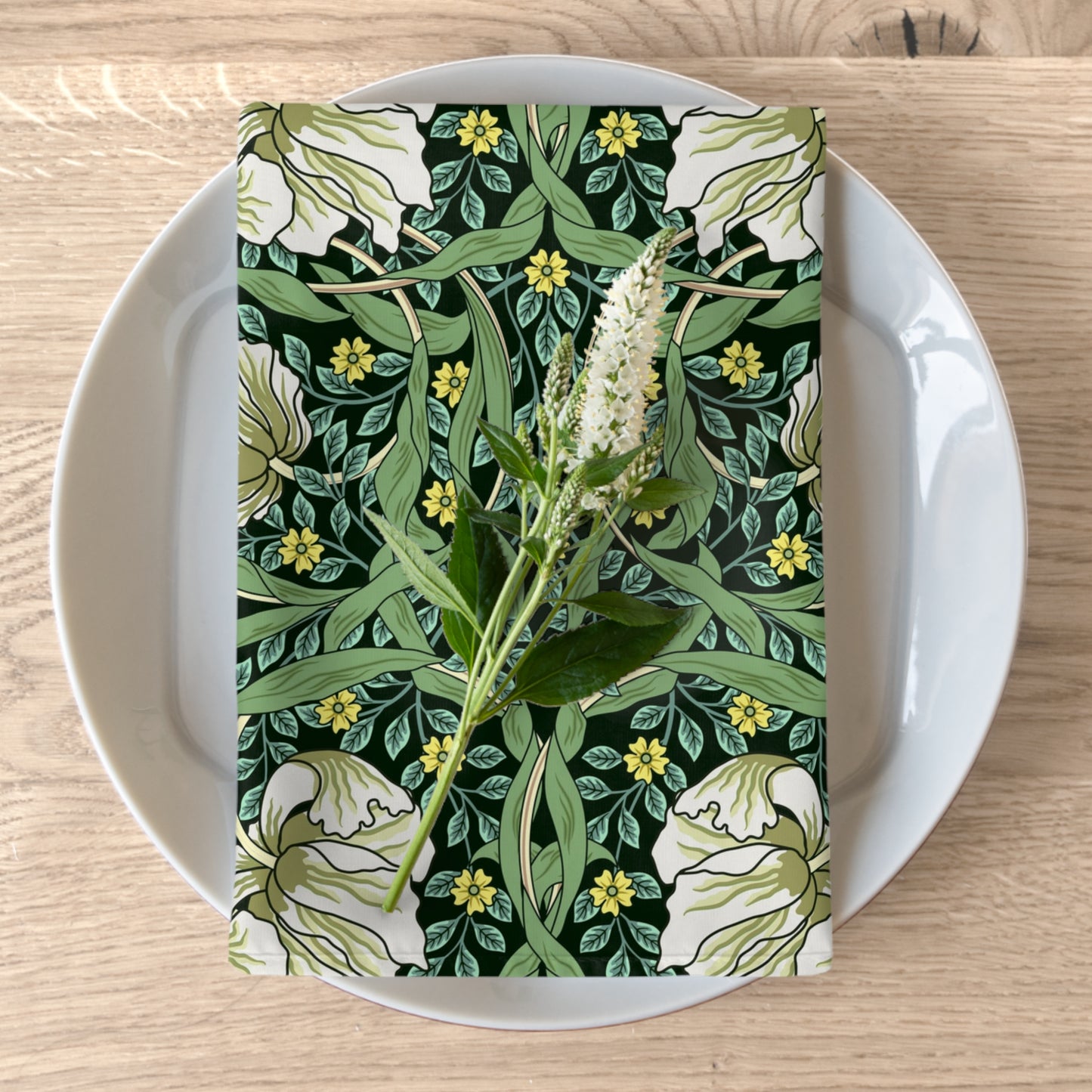 A green floral patterned cotton twill napkin is displayed on a white plate with a piece of floral decor. The napkin has a William Morris inspired design with a variety of botanical motifs.