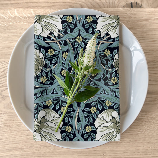 A folded cotton twill napkin with a floral pattern inspired by William Morris, placed on a plate on top of a wooden table.