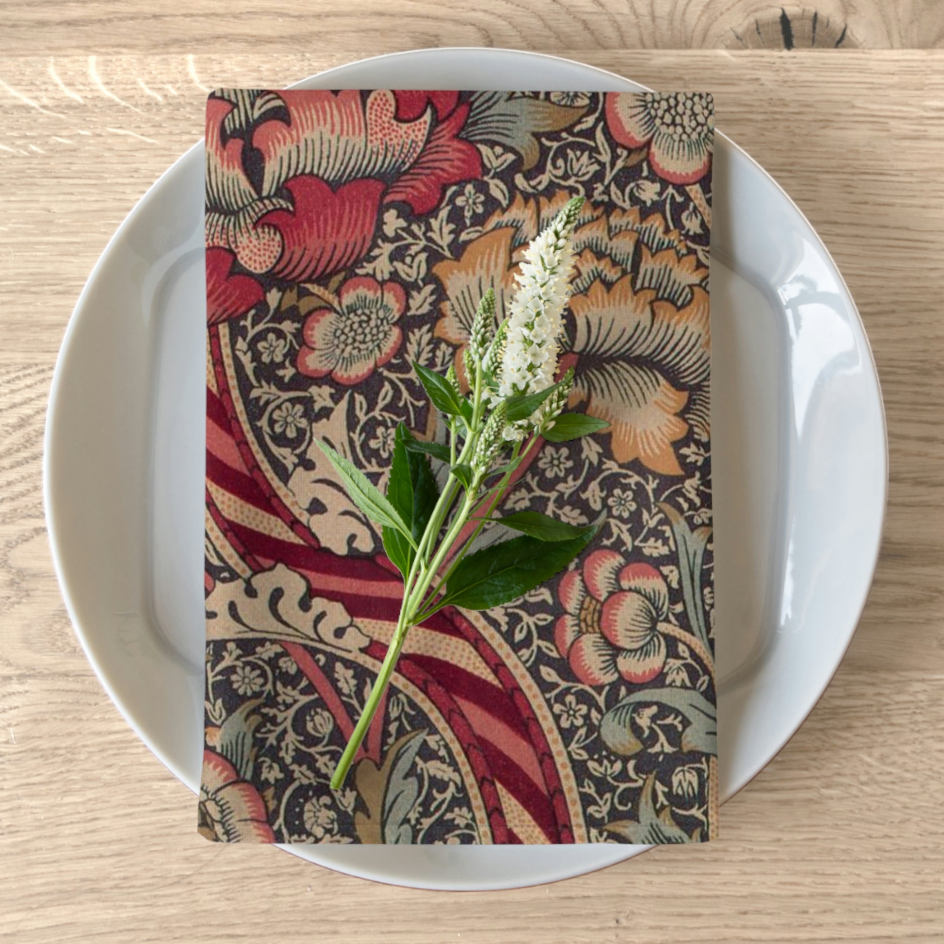 A red floral patterned cotton twill napkin on a white plate with a piece of floral decor on top.