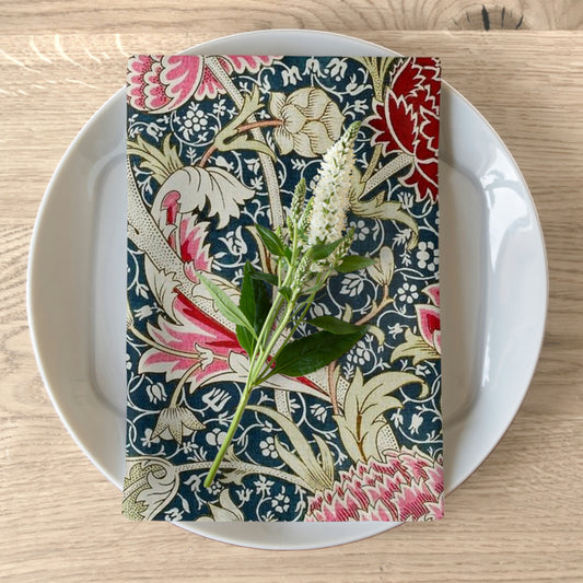 A floral patterned cotton twill napkin inspired by William Morris, displayed on a white plate with a piece of floral decor to the napkin's right.