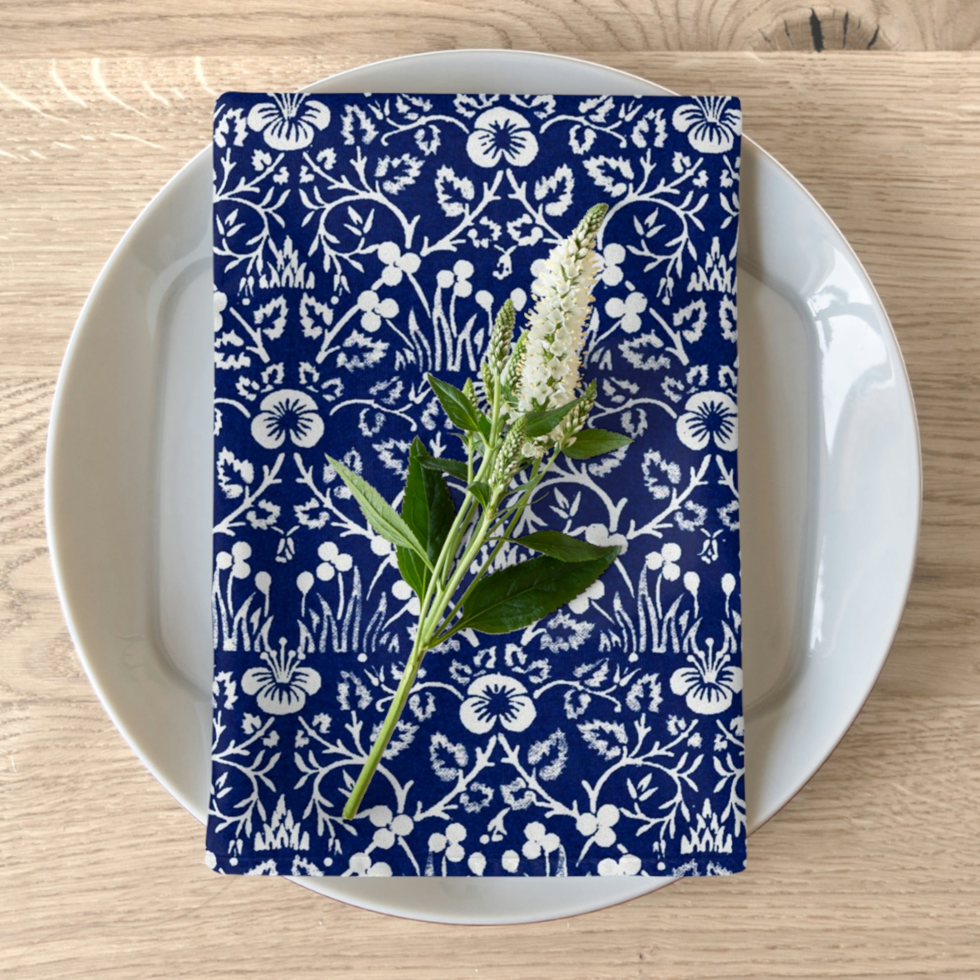 A navy blue cotton twill napkin with a white floral pattern is placed on a white plate on a wooden table. The napkin has hemmed edges and appears soft and elegant.