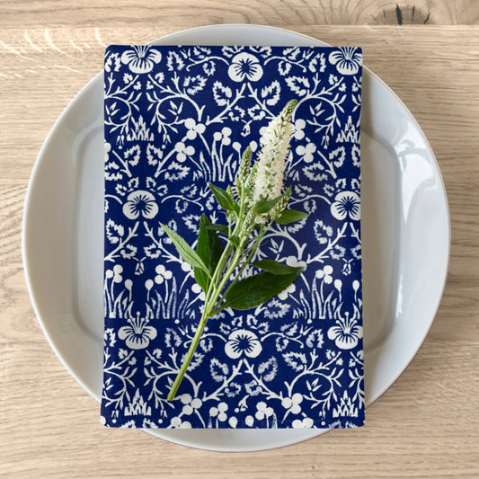 A navy blue cotton twill napkin with a white floral pattern is placed on a white plate on a wooden table. The napkin has hemmed edges and appears soft and elegant.