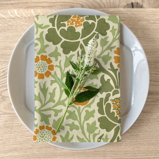 A floral patterned cotton twill napkin is displayed on a plate with a sprig of greenery placed on top, set against a wooden table background.