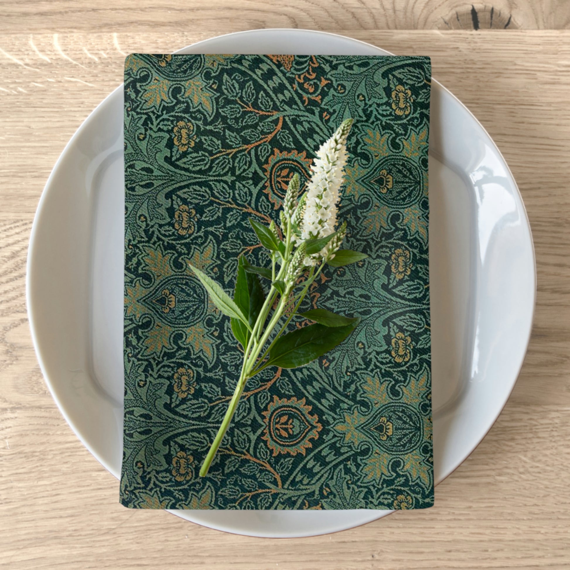 A green cotton twill napkin with a floral pattern is placed on a white plate on a wooden table. The napkin has a hemmed edge and a beautiful floral design.
