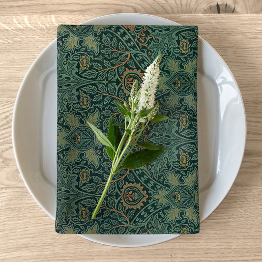 A green cotton twill napkin with a floral pattern is placed on a white plate on a wooden table. The napkin has a hemmed edge and a beautiful floral design.