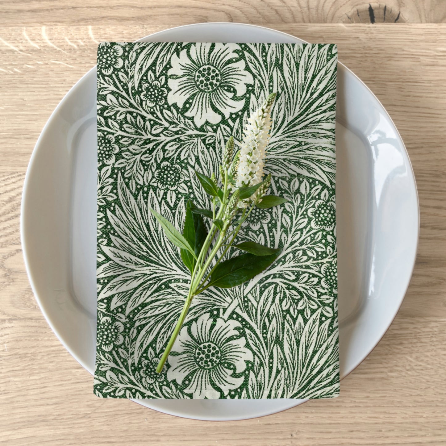 A green floral patterned cotton twill napkin inspired by William Morris, displayed on a white plate with a matching floral arrangement.