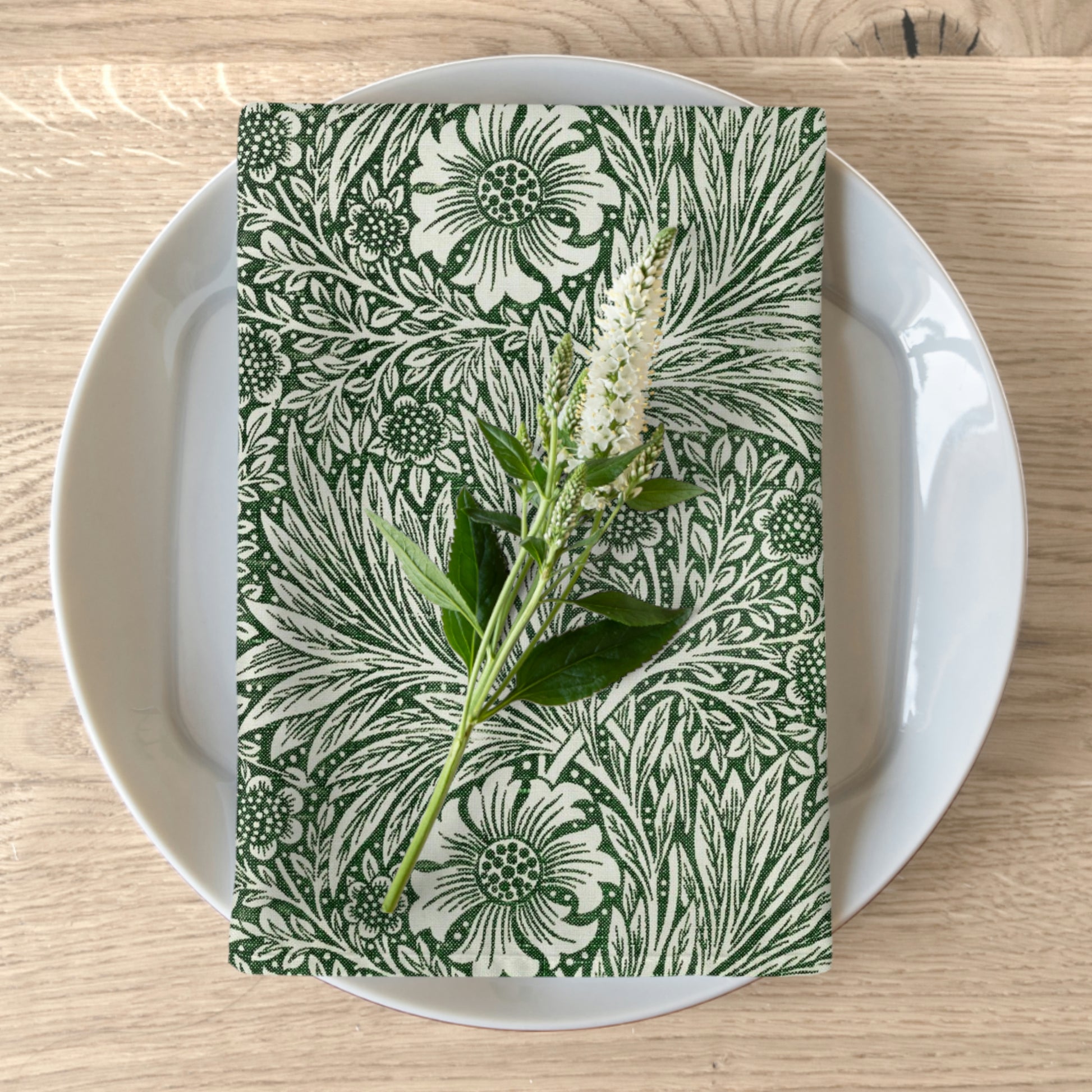 A green floral patterned cotton twill napkin inspired by William Morris, displayed on a white plate with a matching floral arrangement.