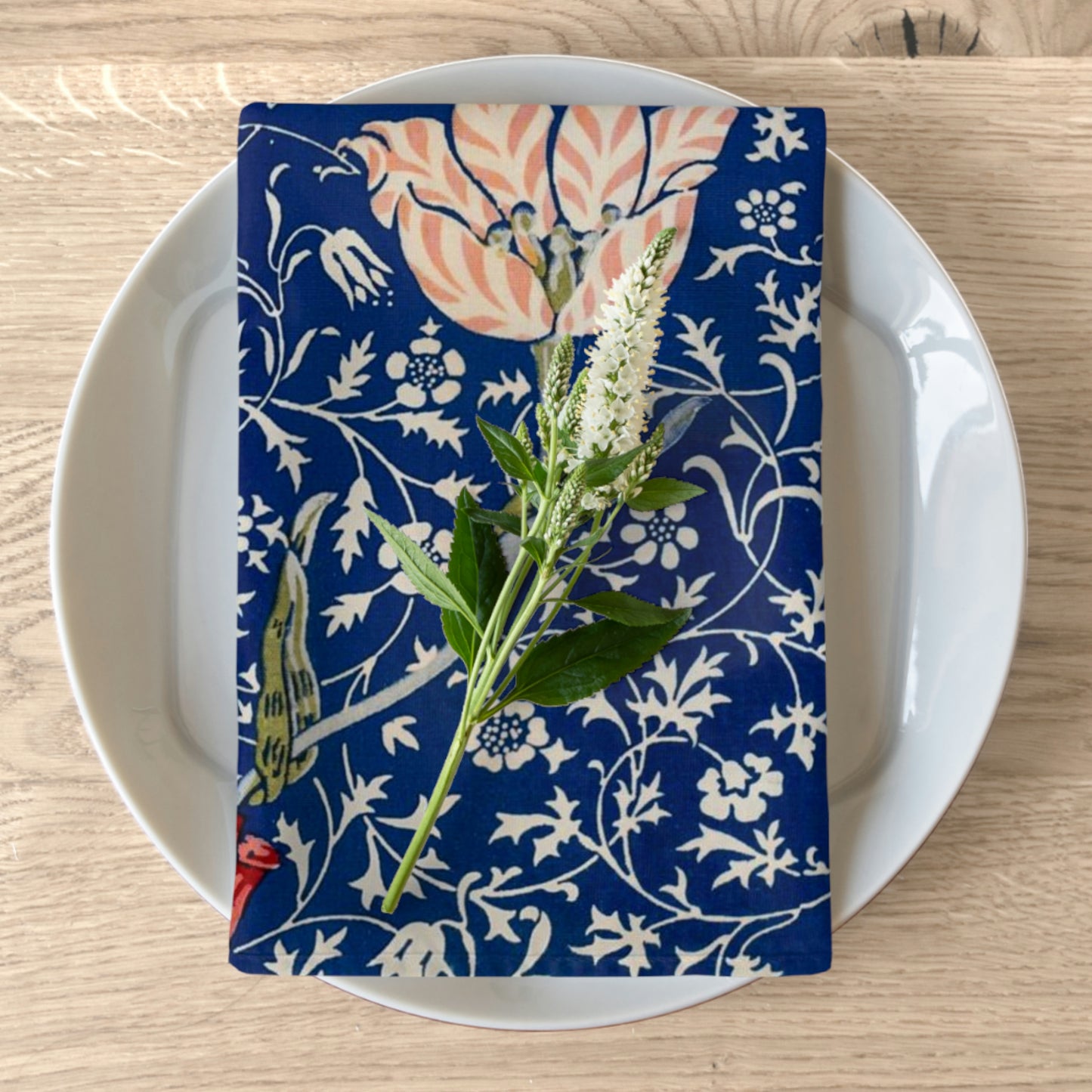 A folded blue cotton twill napkin with a floral pattern inspired by William Morris, displayed on a white plate on a wooden table.