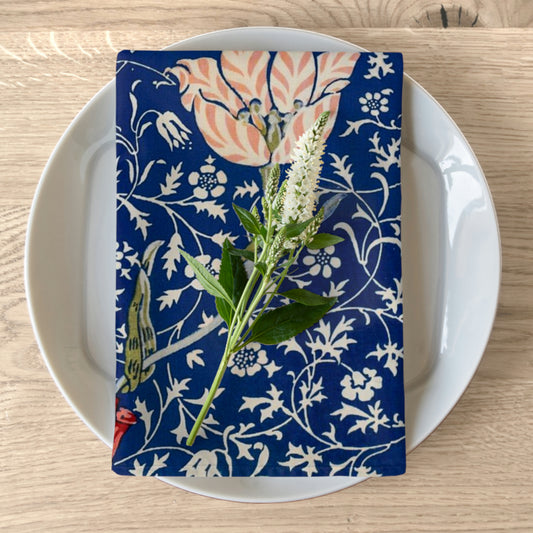 A folded blue cotton twill napkin with a floral pattern inspired by William Morris, displayed on a white plate on a wooden table.