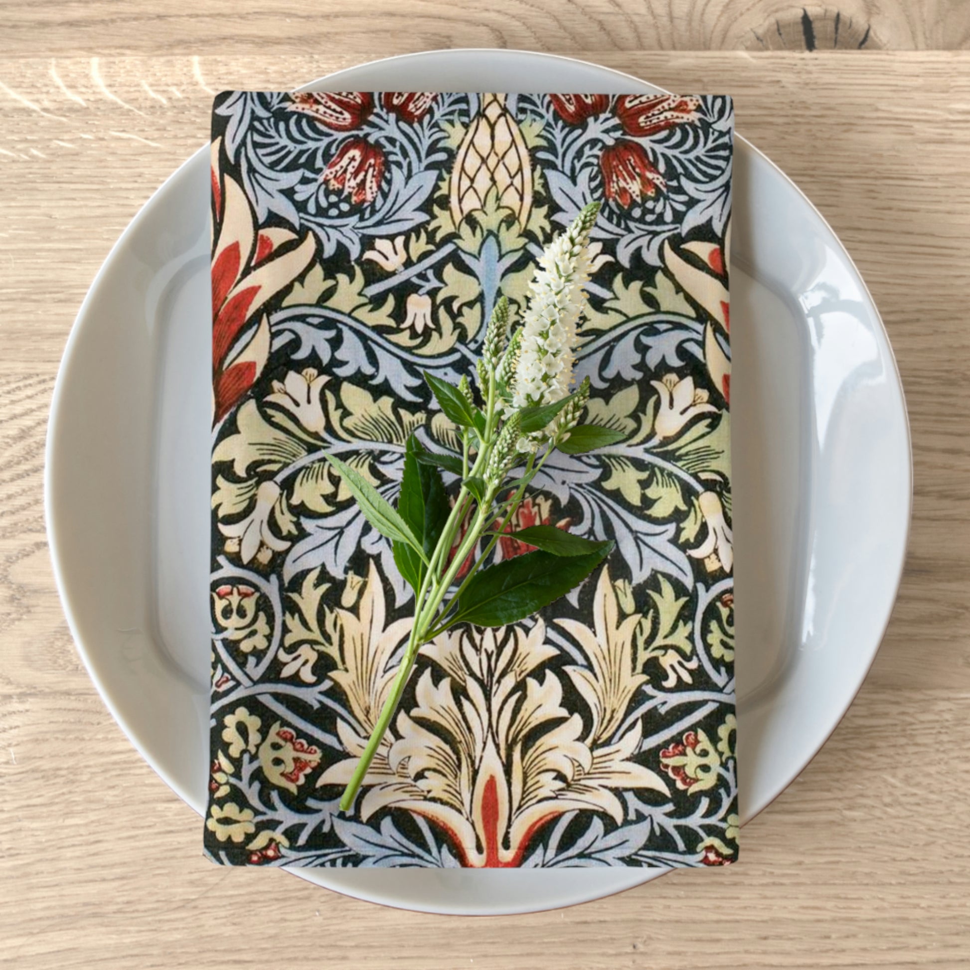 A floral patterned cotton twill napkin inspired by William Morris, displayed on a plate with a white flower decoration.