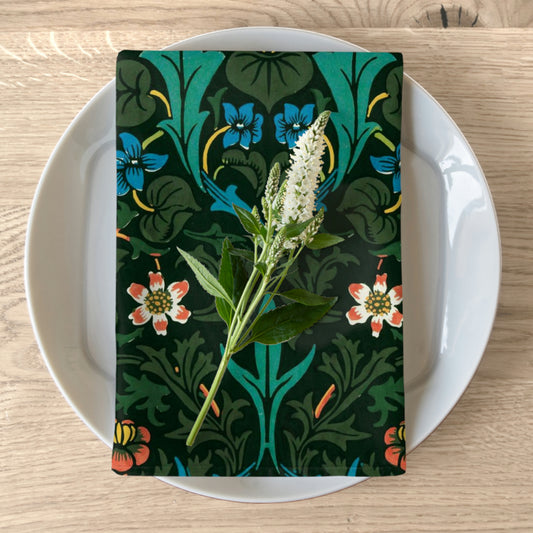 A floral patterned cotton twill napkin inspired by William Morris, displayed on a plate with a white flower decoration.