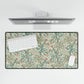 A desk mat with a floral pattern, inspired by William Morris, displayed under a desk with a keyboard and mouse on top.