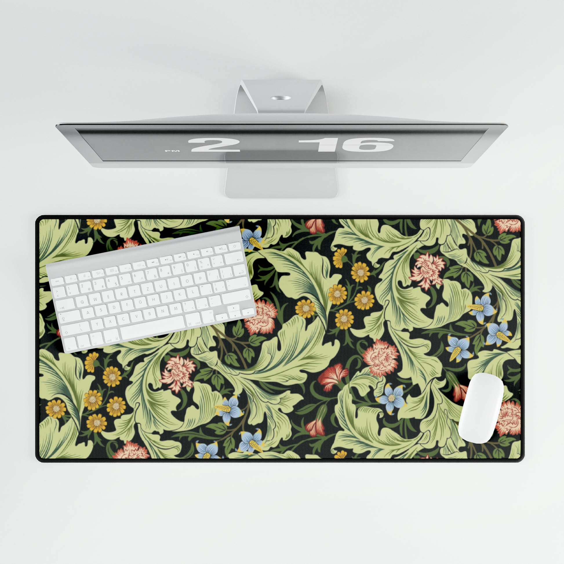 A large desk mat with a green William Morris & Co floral pattern, displayed on a desk with a keyboard and mouse, with a monitor above it.