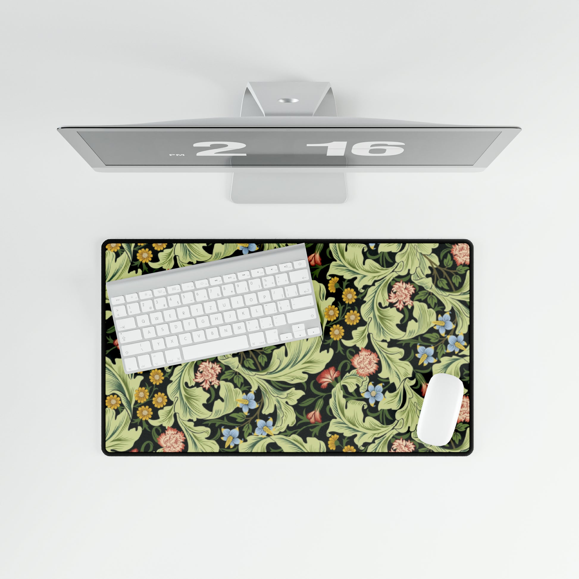 A large desk mat with a green William Morris & Co floral pattern, displayed on a desk with a keyboard and mouse, with a monitor above it.
