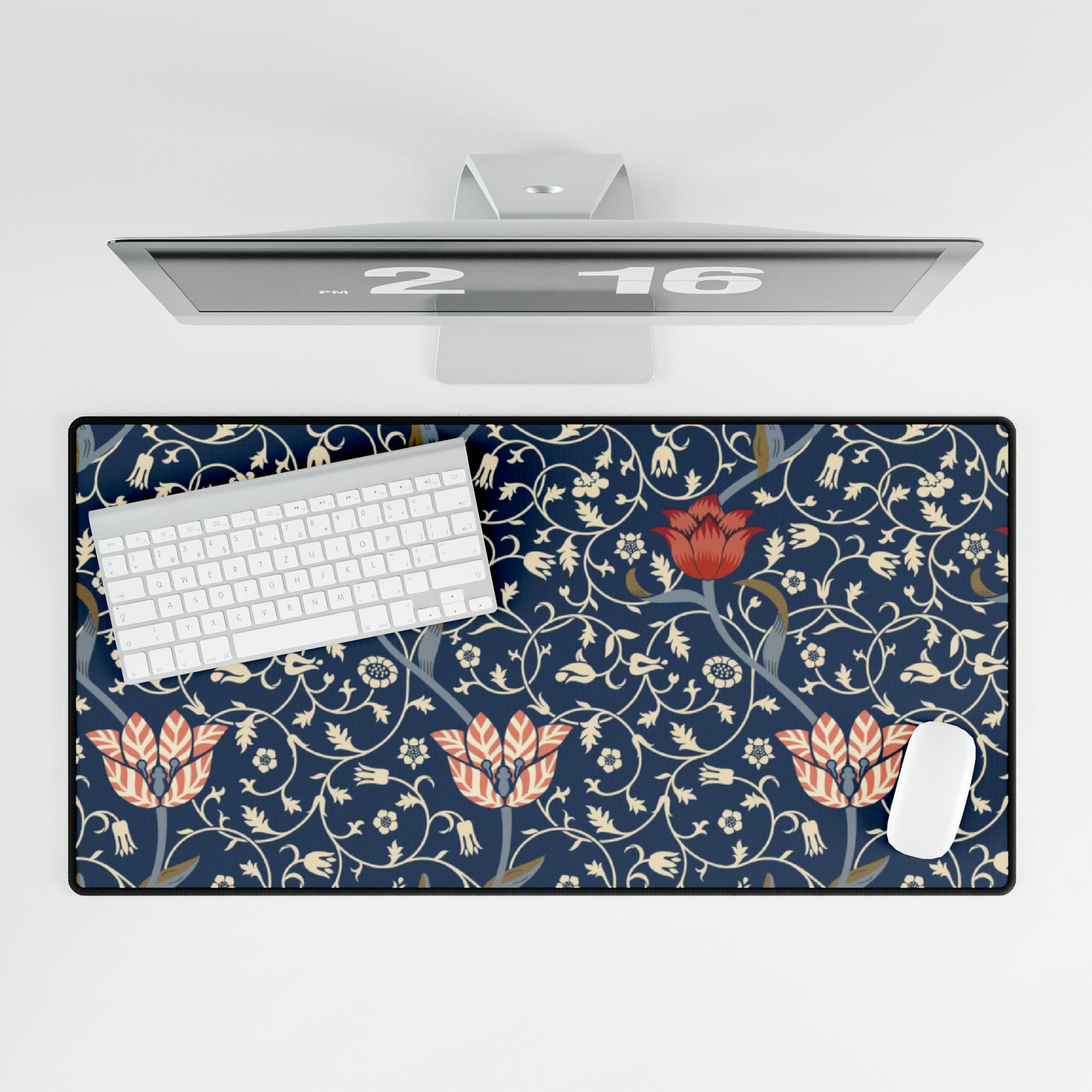 A large desk mat with a floral pattern in shades of red, pink and blue on a dark background, displayed under a monitor with a keyboard and mouse on top.