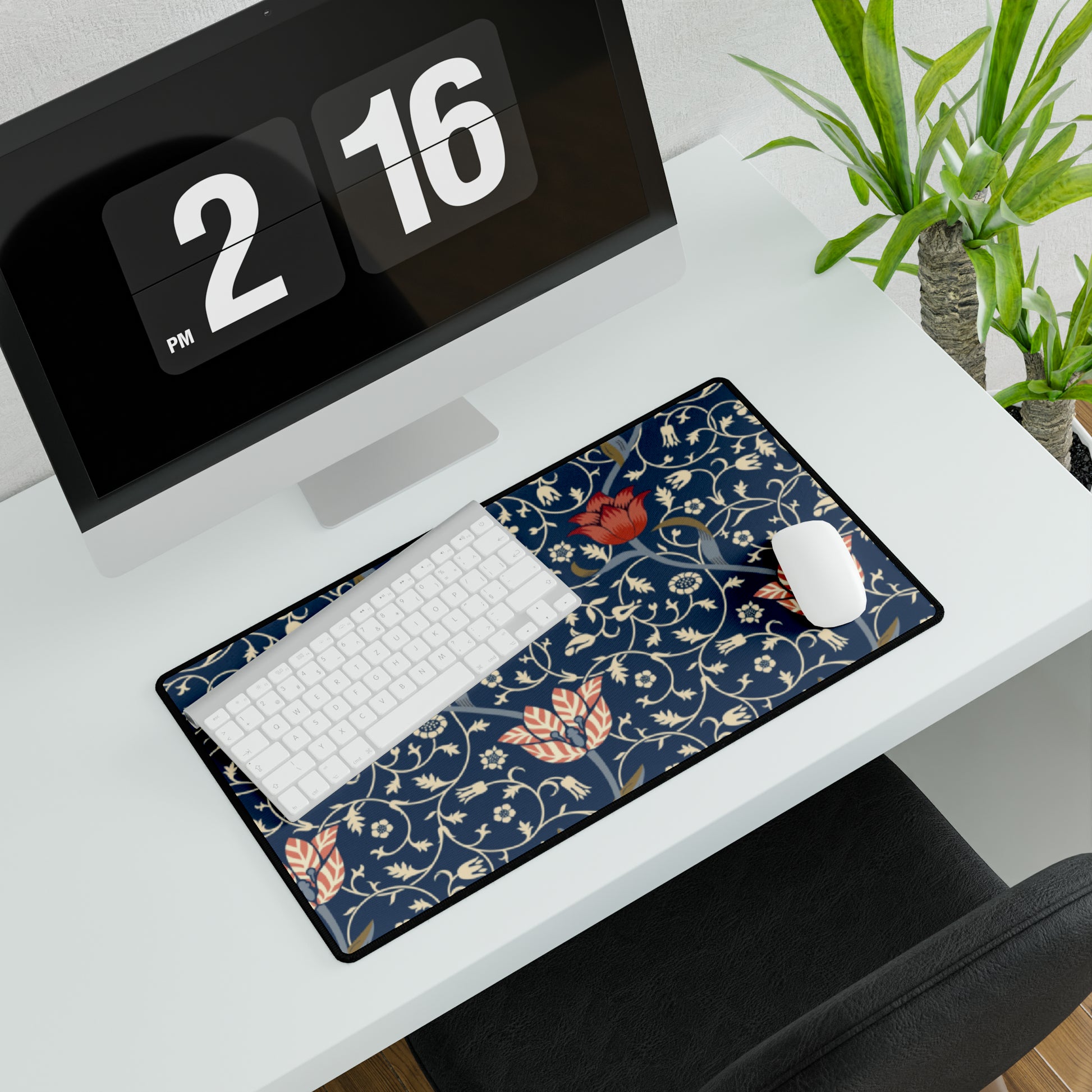 A large desk mat with a floral pattern in shades of red, pink and blue on a dark background, displayed under a monitor with a keyboard and mouse on top.
