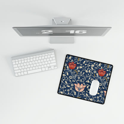 A large desk mat with a floral pattern in shades of red, pink and blue on a dark background, displayed under a monitor with a keyboard and mouse on top.
