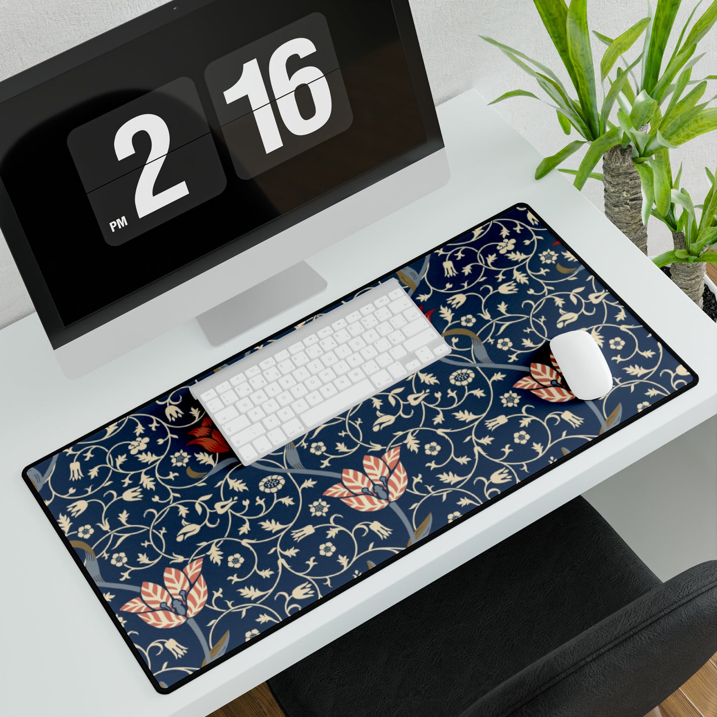 A large desk mat with a floral pattern in shades of red, pink and blue on a dark background, displayed under a monitor with a keyboard and mouse on top.