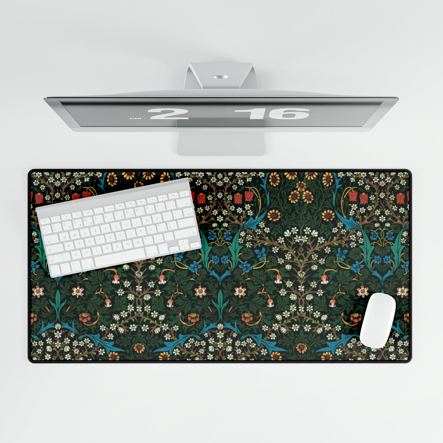 A rectangular desk mat with a William Morris & Co floral tulip pattern in red, black, and green, placed under a white desk lamp and a white keyboard.