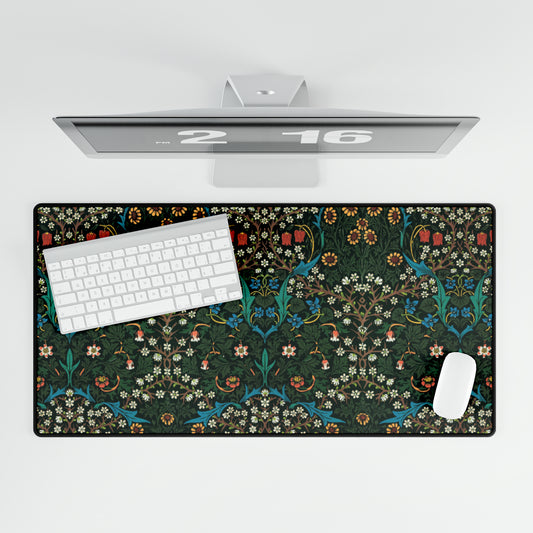 A rectangular desk mat with a William Morris & Co floral tulip pattern in red, black, and green, placed under a white desk lamp and a white keyboard.