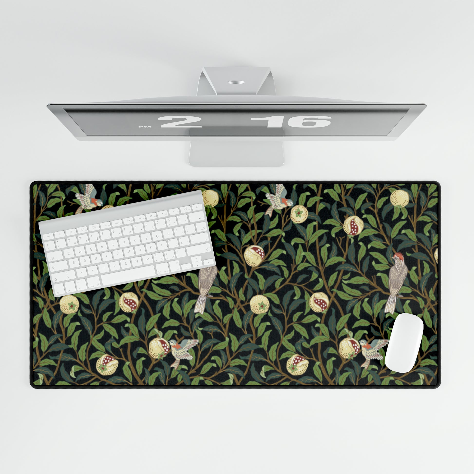 A rectangular desk mat with a floral William Morris & Co pattern featuring birds and pomegranates, placed under a desk with a keyboard and mouse on top.