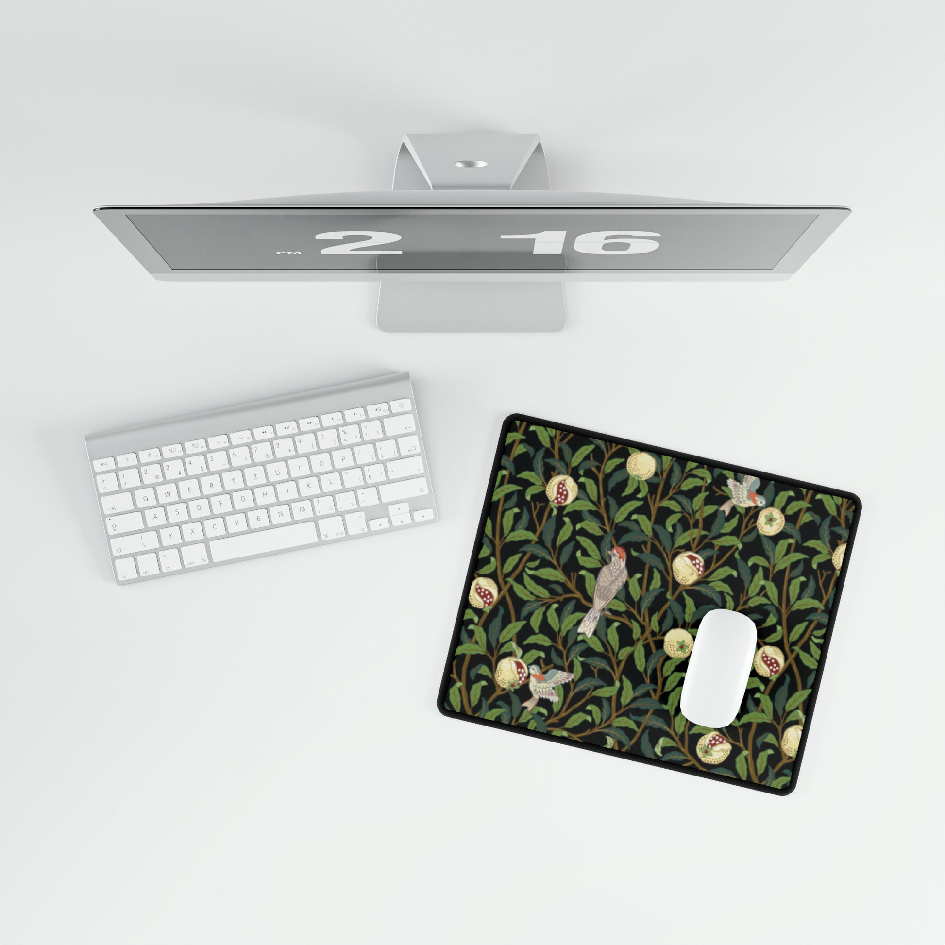 A rectangular desk mat with a floral William Morris & Co pattern featuring birds and pomegranates, placed under a desk with a keyboard and mouse on top.