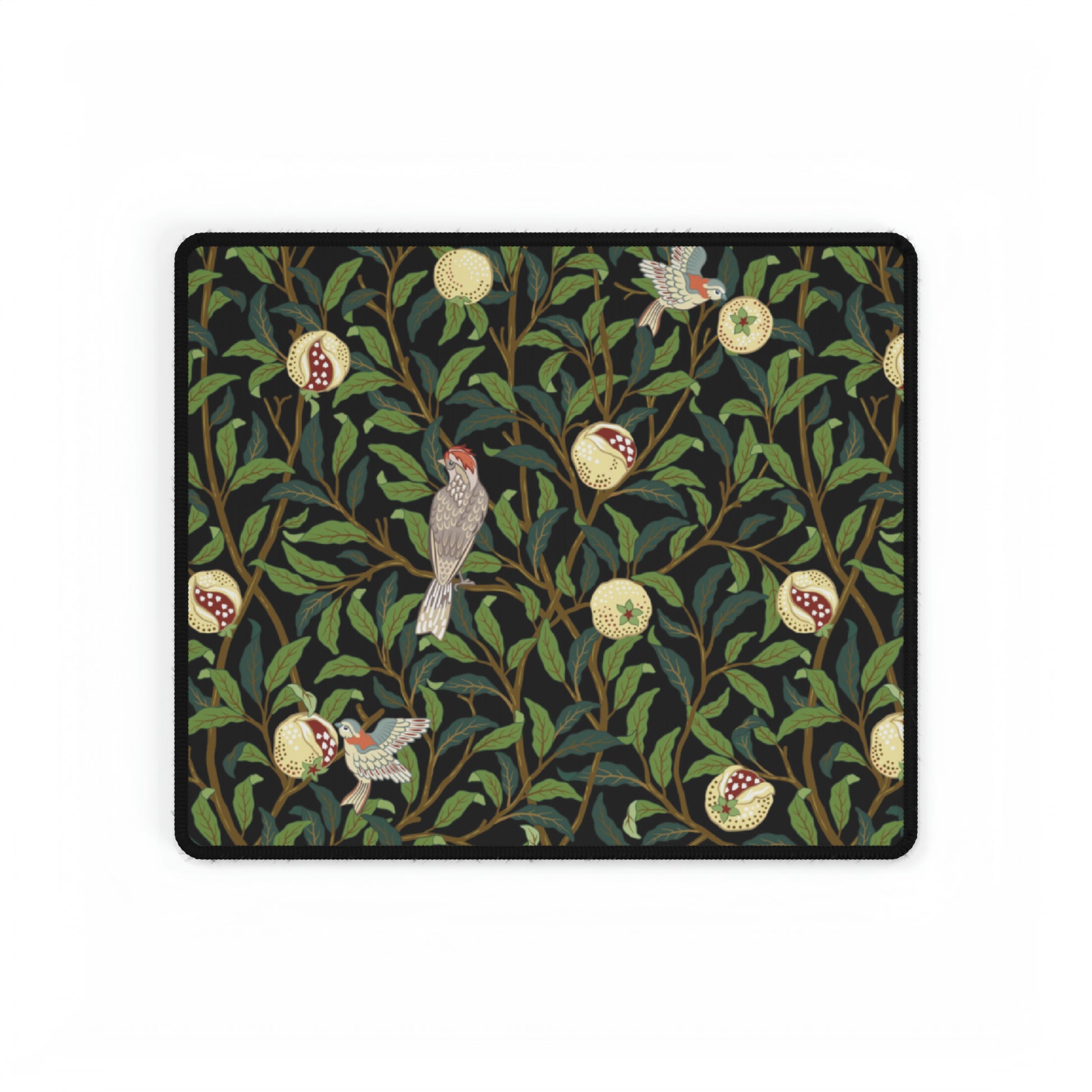A rectangular desk mat with a floral William Morris & Co pattern featuring birds and pomegranates, placed under a desk with a keyboard and mouse on top.