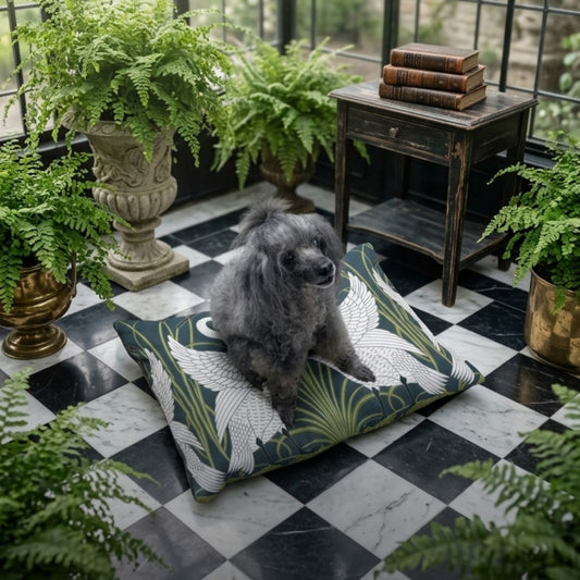 A small gray dog sits on a large decorative floor cushion with a green, white, and beige tropical leaf pattern, placed on a black-and-white checkered balcony floor surrounded by potted ferns.